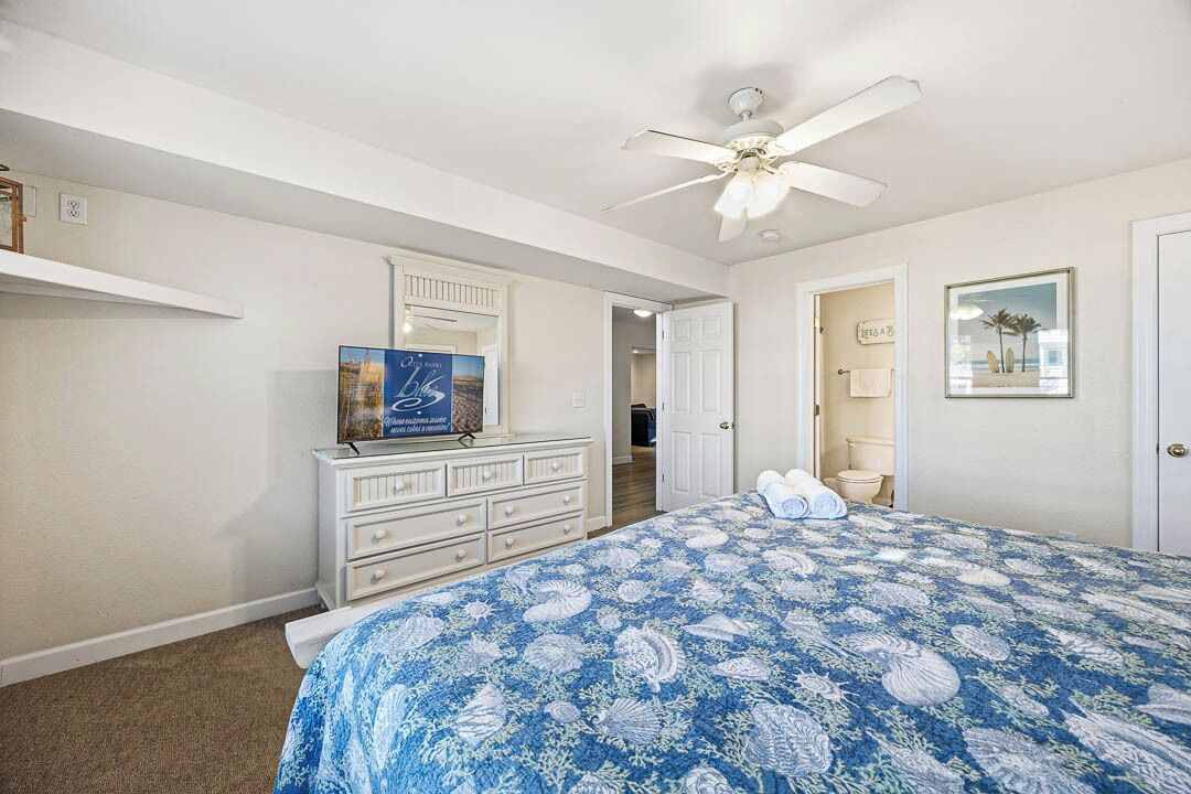 A bedroom with a bed, a dresser, a ceiling fan, and a door leading to another room.