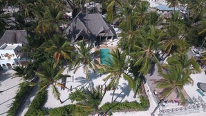 Vista aérea de um resort à beira-mar com edifícios de telhado de palha, palmeiras e uma piscina.