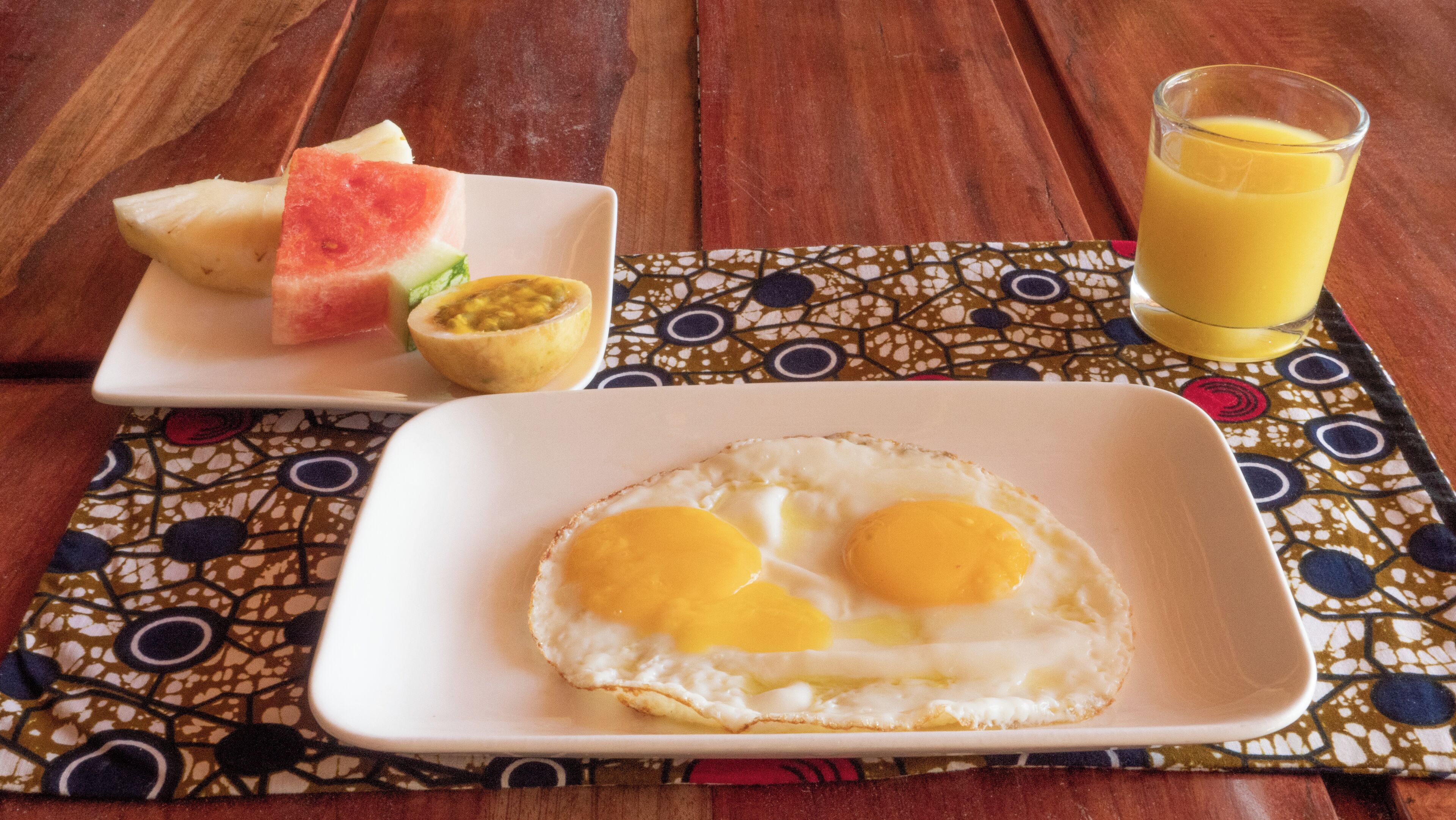 A breakfast plate with eggs, a slice of watermelon, and a bowl of fruit salad on a patterned placemat.