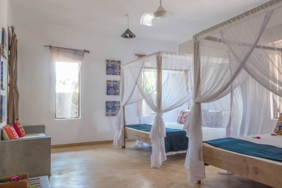 Habitación con tres camas, cada una con dosel. Hay tres unidades de aire acondicionado fijadas a la pared, una ventana con cortinas translúcidas y piso de concreto.