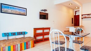 Uma área de refeições com mesa e cadeiras, um banco colorido, uma TV fixada na parede e um letreiro escrito 'PARA A PRAIA'.