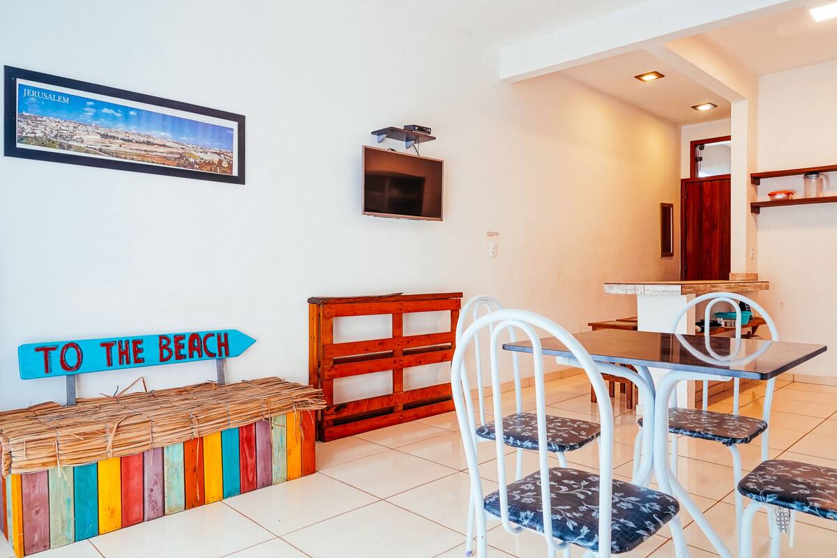 A dining area with a table and chairs, a colorful bench, a wall-mounted TV, and a 'TO THE BEACH' sign.