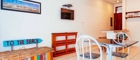 A dining area with a table and chairs, a colorful bench, a wall-mounted TV, and a 'TO THE BEACH' sign.