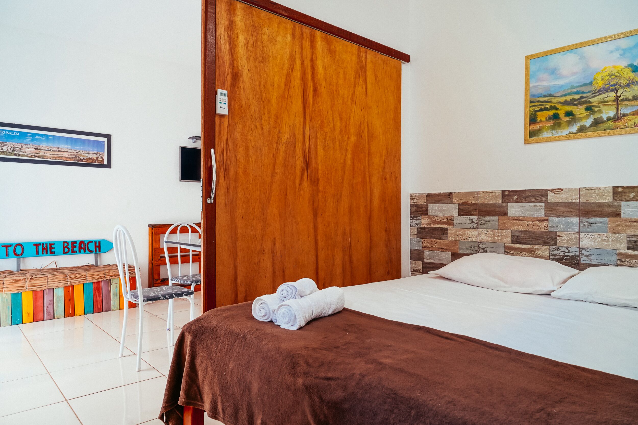 Ein Schlafzimmer mit einem Holzschrank, einem Bett mit brauner Decke, einem Bild an der Wand und einem Schild mit der Aufschrift 'TO THE BEACH'.