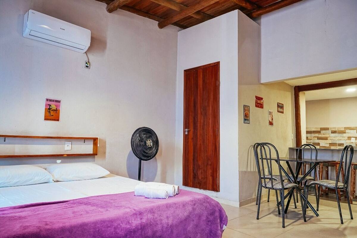 A bedroom with a bed, a fan, a wooden door, and a dining area with chairs and a table.