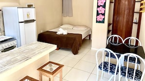 Quarto pequeno e limpo com cama, mesa, cadeiras, frigorífico, fogão e porta.