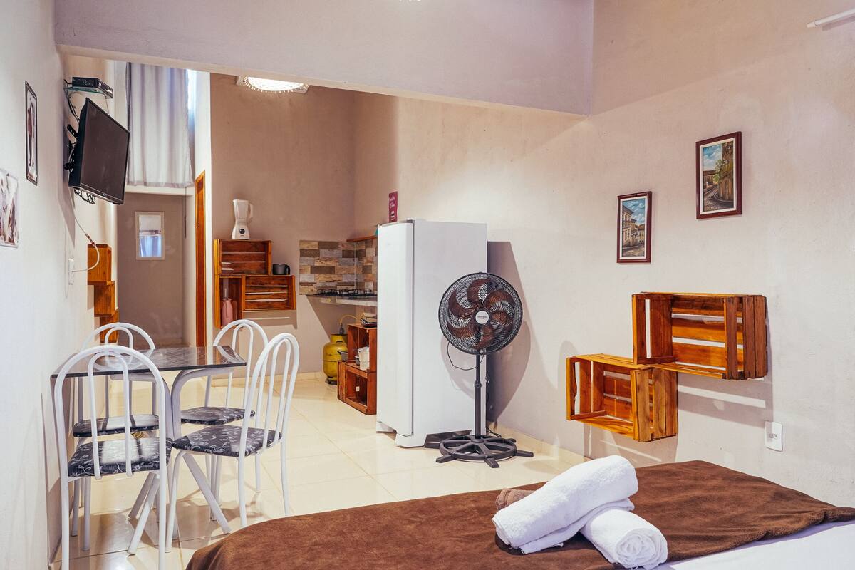 A compact room with a bed, a dining area with chairs, a small kitchen with a refrigerator, and a fan.