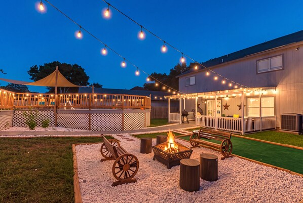 An outdoor patio area with string lights, a fire pit, wooden benches, and a covered seating area.