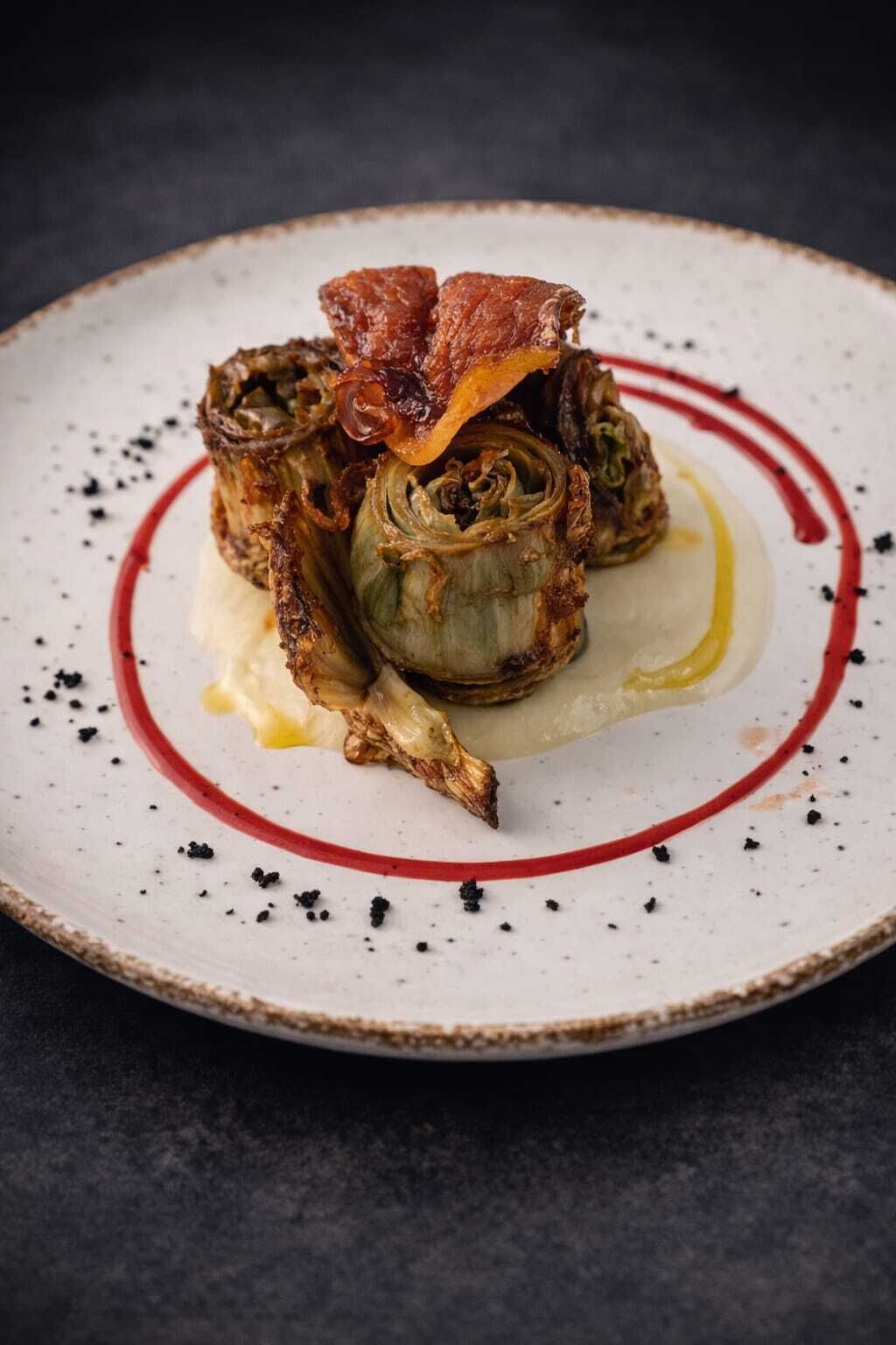 A plate with a dish of artichoke hearts and bacon, garnished with black pepper and a red sauce.
