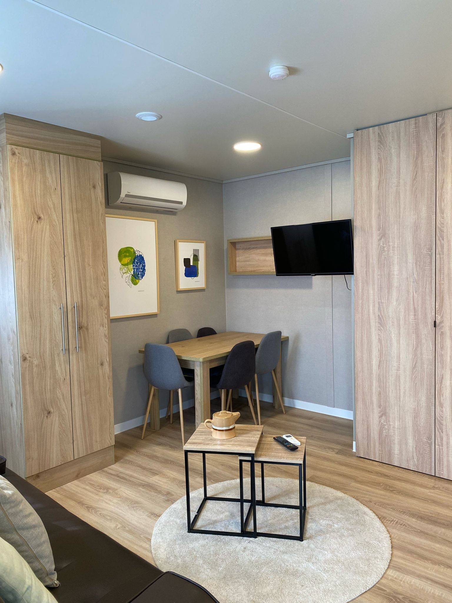Una sala moderna con una mesa de comedor de madera, sillas grises, un televisor montado en la pared y un armario grande.