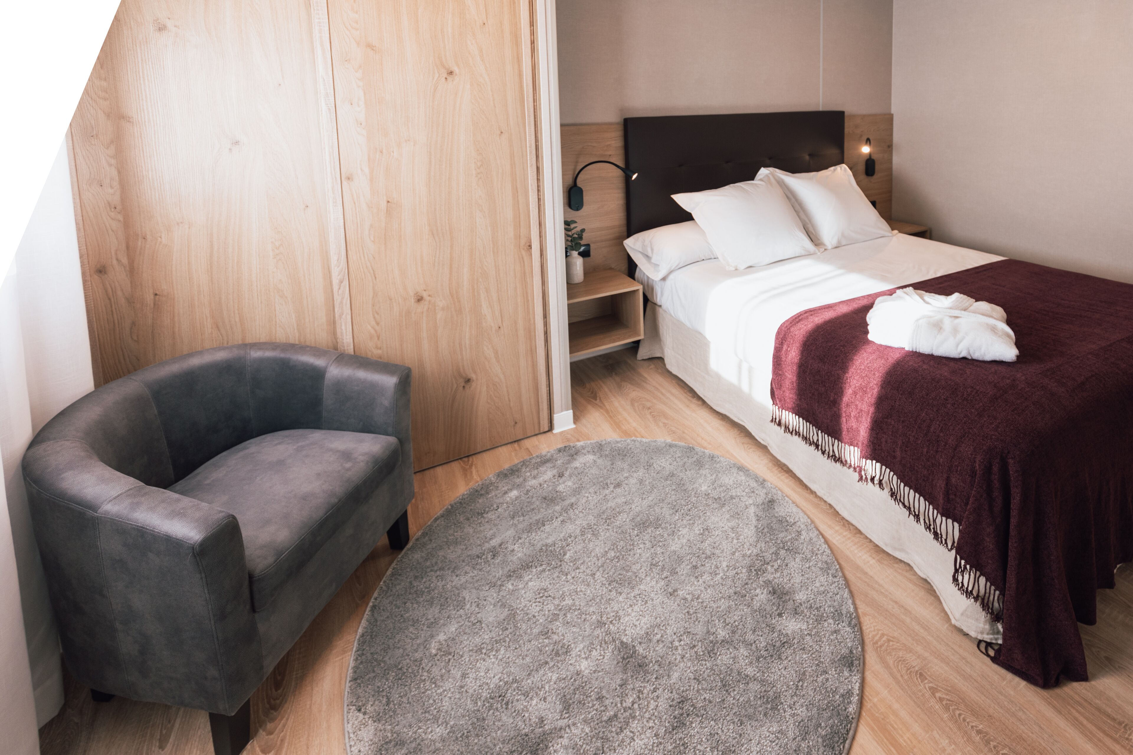 A modern hotel room with a bed, a grey armchair, a round grey rug, and a wooden wardrobe.