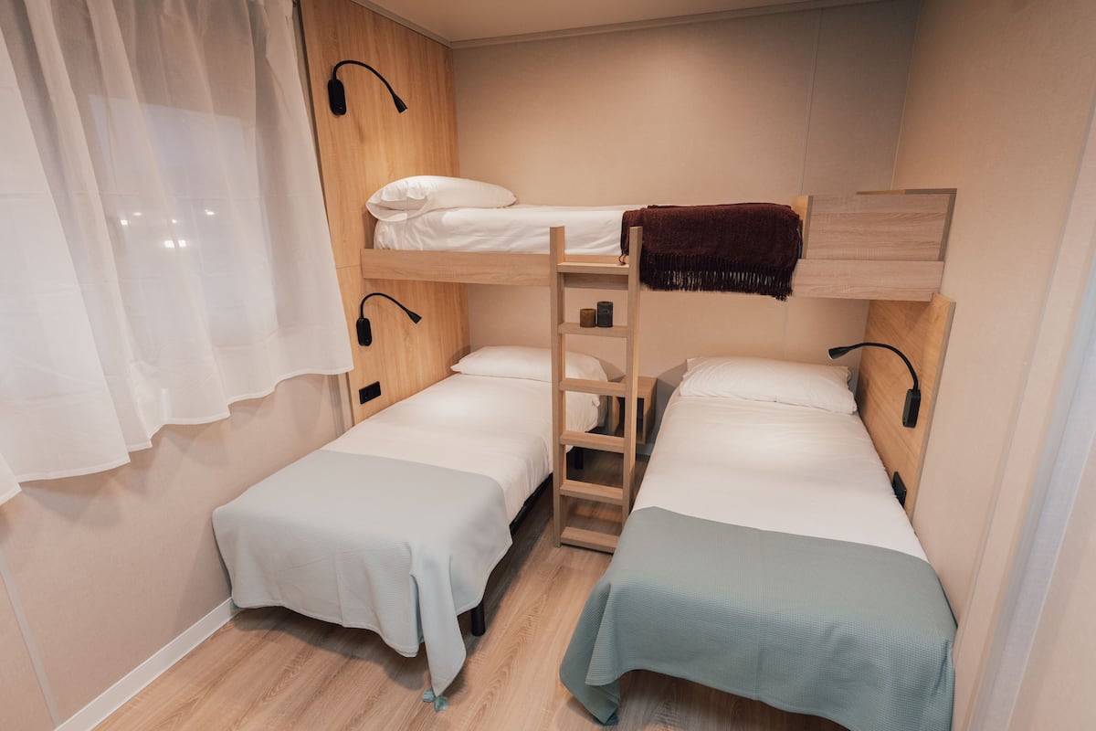 A compact room with two single beds, a bunk bed, a wooden ladder, and a white curtain.