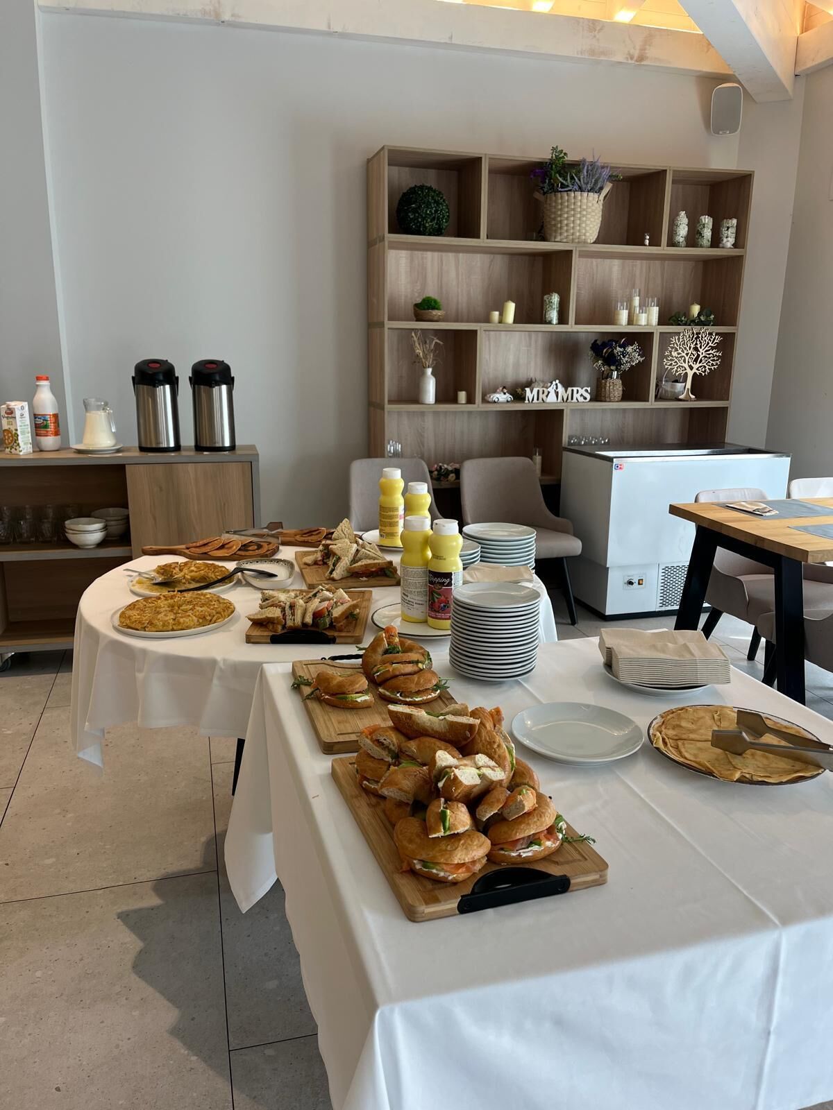 A buffet setup with various food items, including sandwiches and a pie, on tables covered with white tablecloths. There are condiment bottles, plates, and a refrigerator in the background.