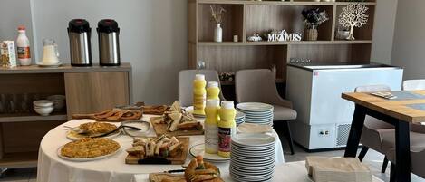 A buffet setup with various food items, including sandwiches and a pie, on tables covered with white tablecloths. There are condiment bottles, plates, and a refrigerator in the background.