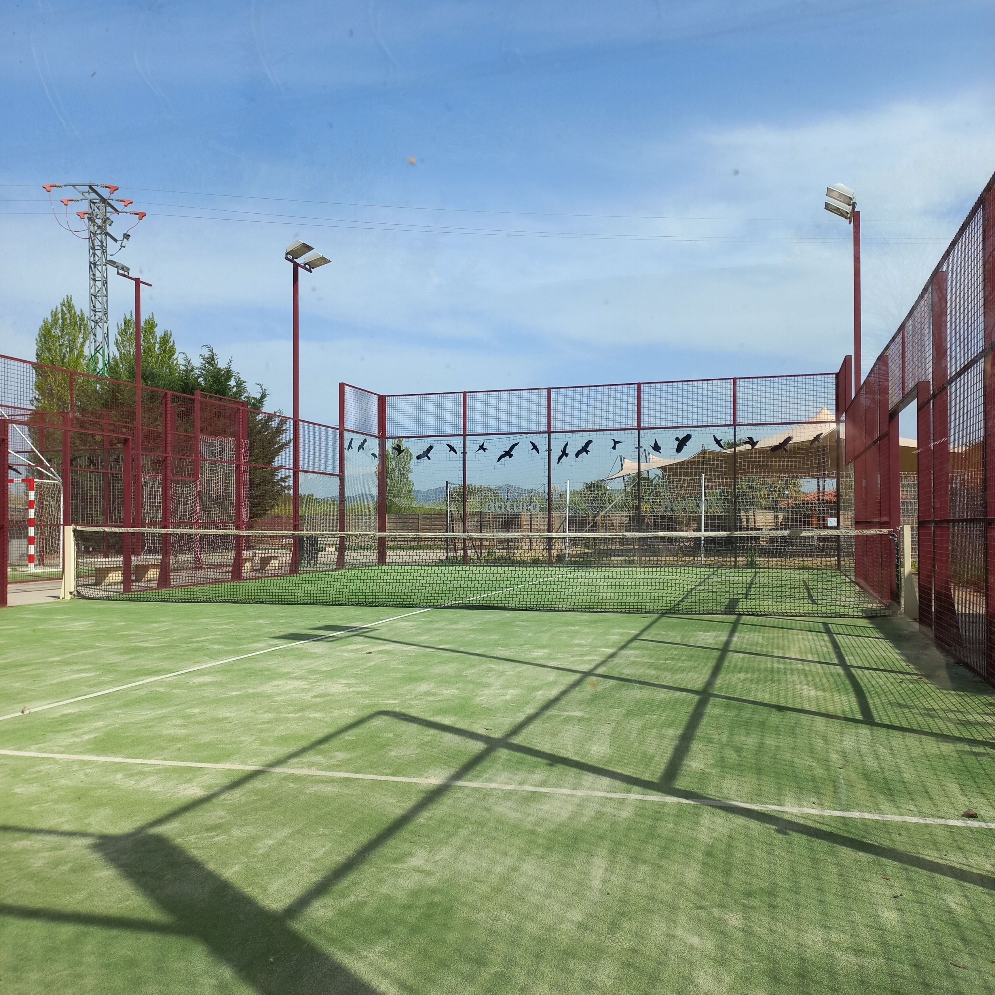 Una cancha de tenis con una red, rodeada por una valla roja y equipada con iluminación artificial.