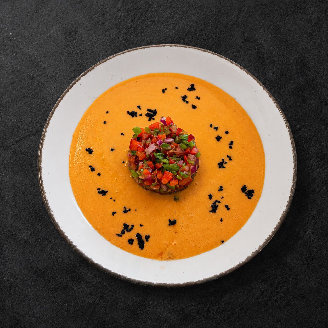 A plate with a central mound of diced vegetables on a smooth orange puree, surrounded by a white rimmed bowl on a dark surface.