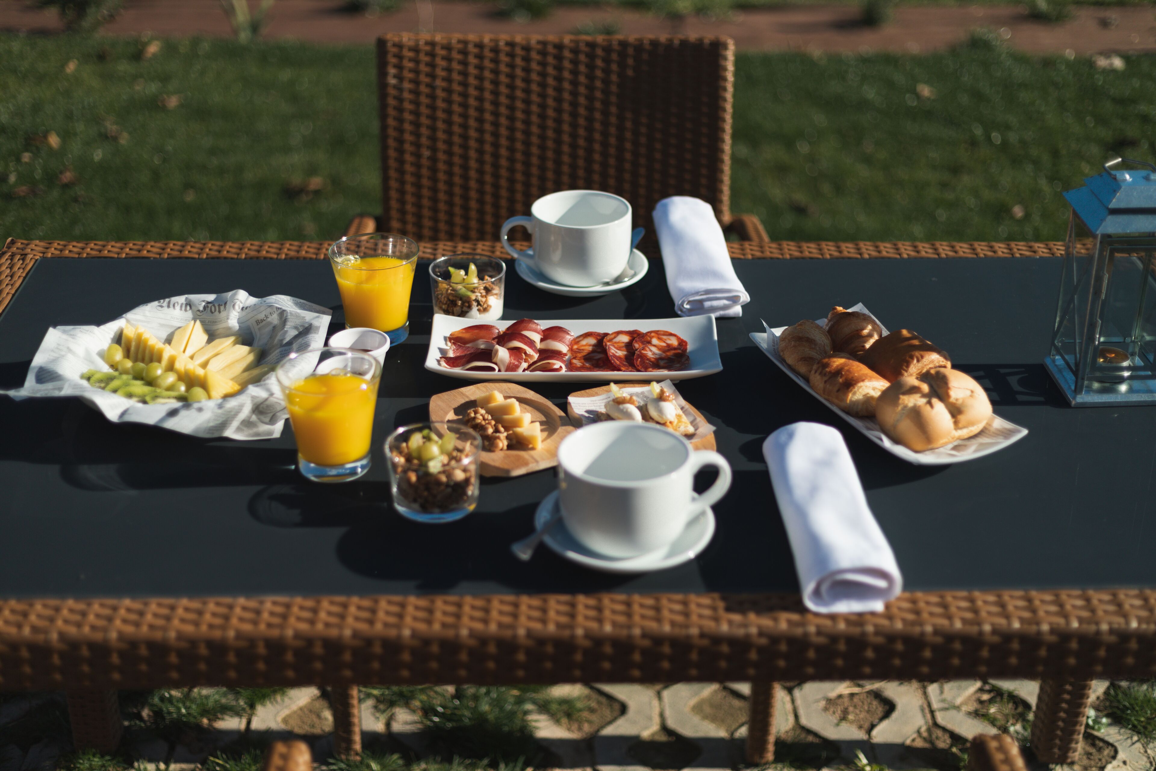 A breakfast table with various food items and drinks, set outdoors on a wicker table.
