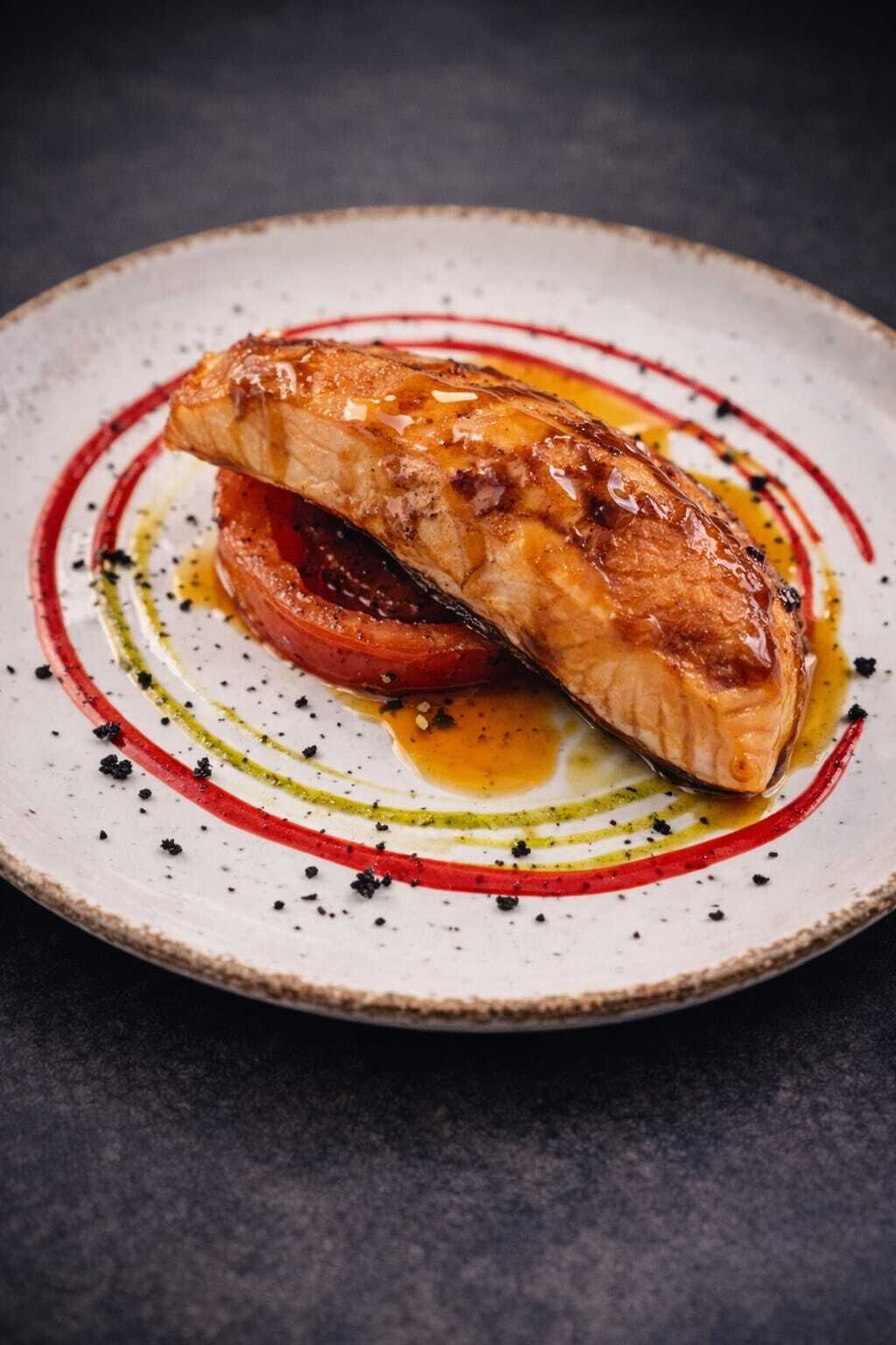 A plate with a piece of grilled fish, a slice of tomato, and a decorative sauce pattern.