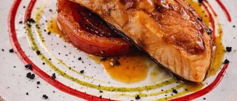 A plate with a piece of grilled fish, a slice of tomato, and a decorative sauce pattern.