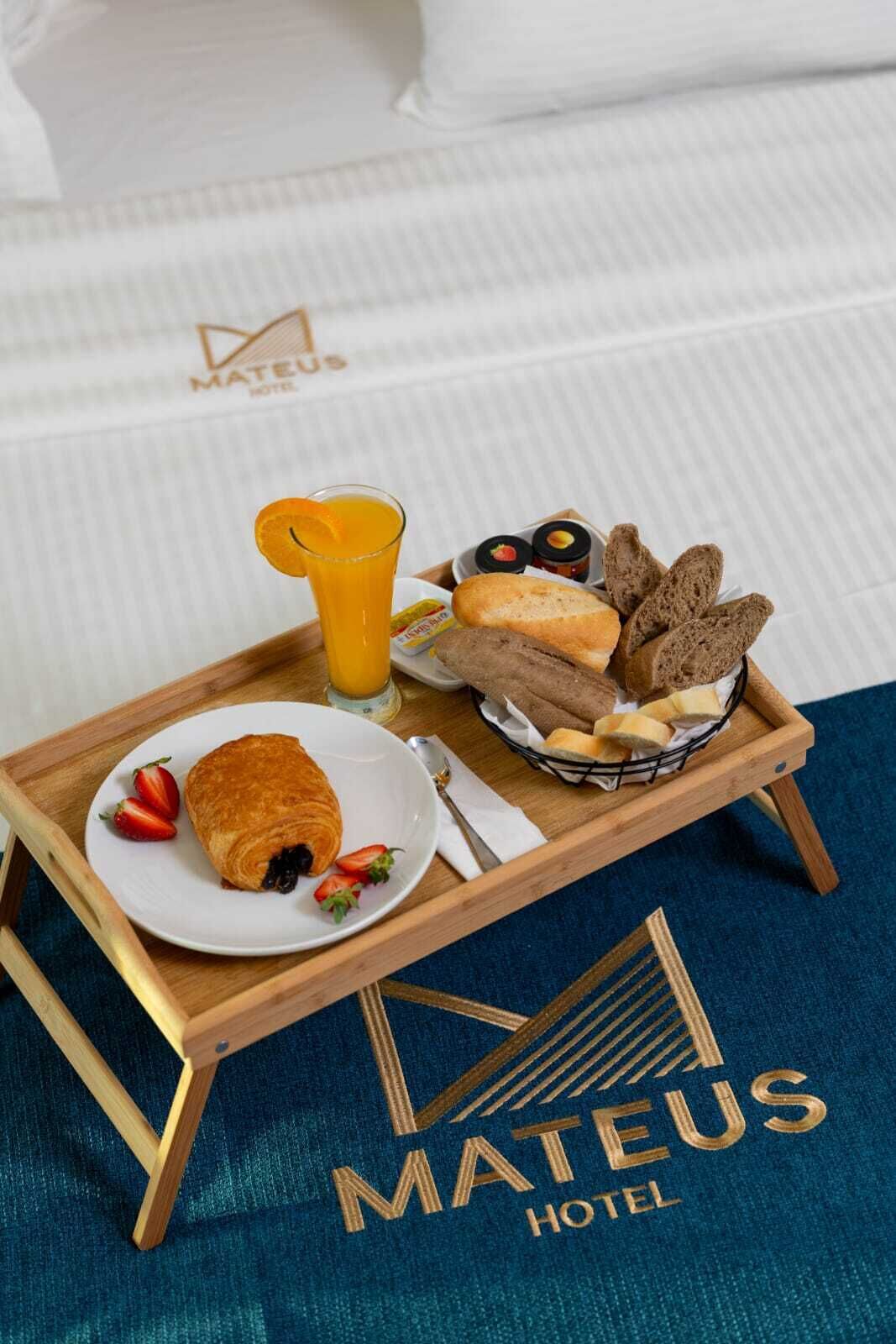 Un plateau de petit-déjeuner avec des croissants, des fraises, du jus d’orange, du pain et des confitures, posé sur un lit recouvert d’une couverture bleue et accompagné d’un plateau en bois.