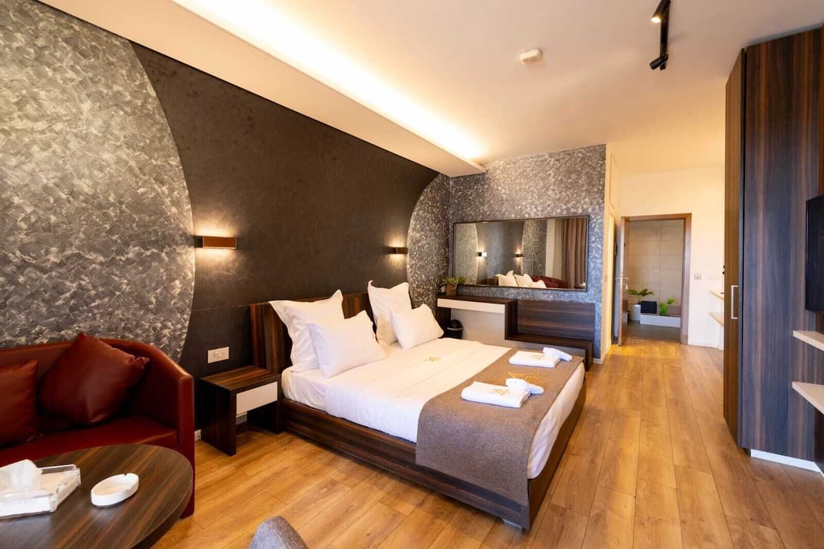 A modern hotel room with a large bed, a red sofa, a wooden floor, a mirror, and a TV.