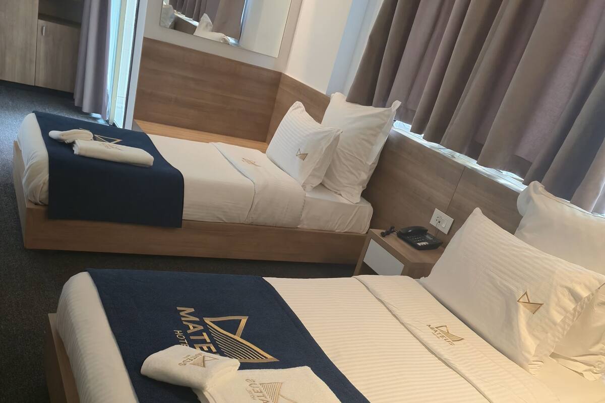 A hotel room with two beds, each with a blue towel and a white pillow with a logo. There is a wooden nightstand with a phone and a small table.