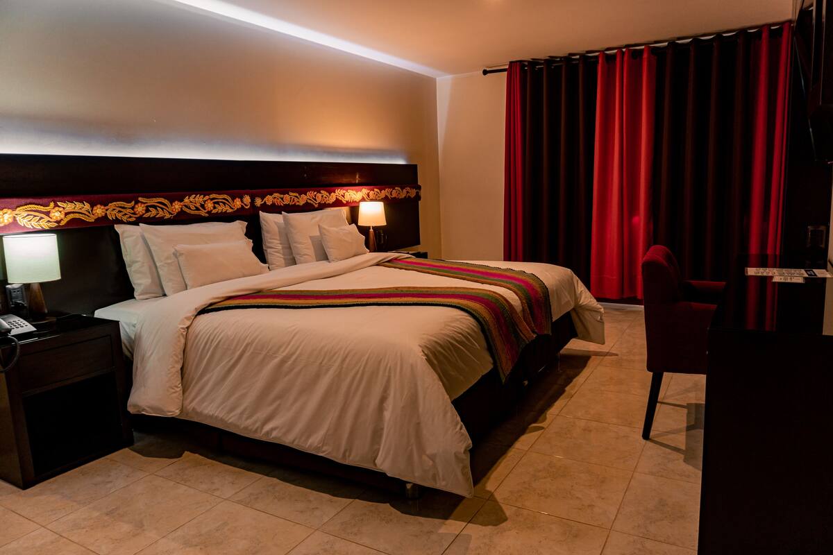 A hotel room with a large bed, a desk with a lamp, a chair, and a red curtain.