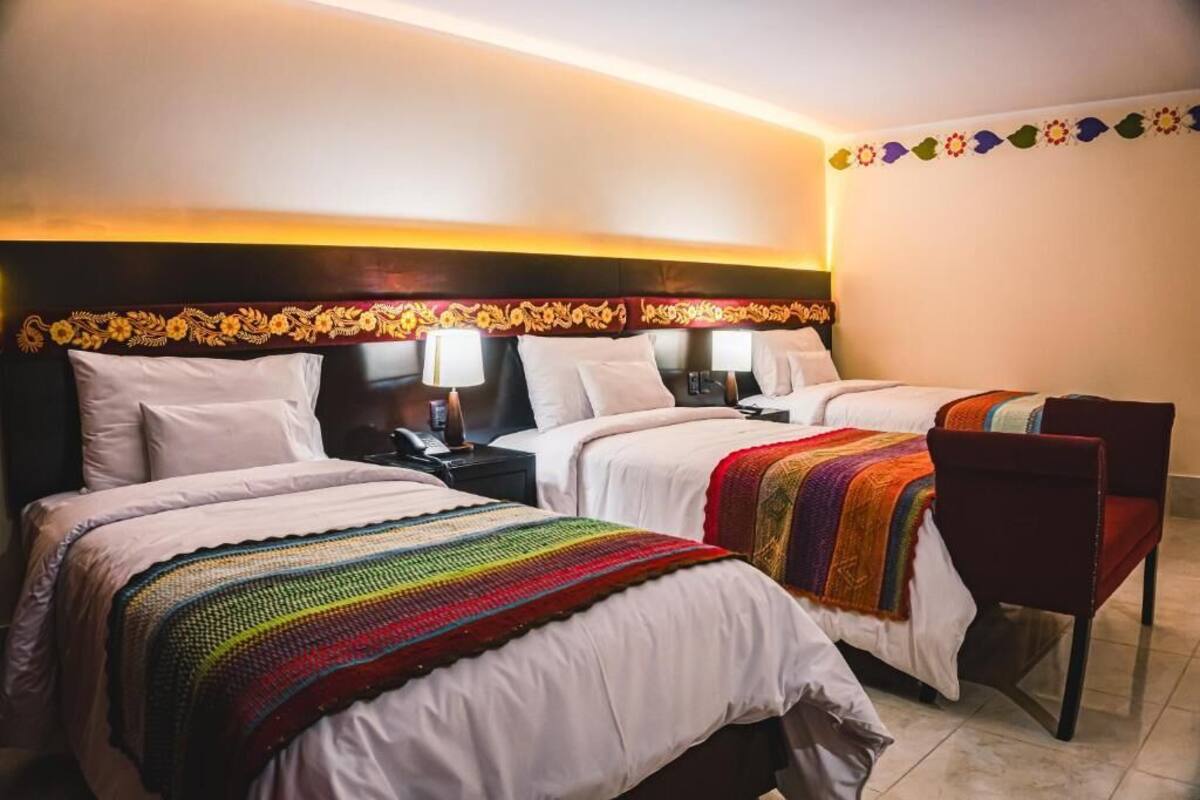 A hotel room with three beds, each with white bedding and a colorful blanket. There are two bedside tables with lamps, and a chair is visible in the corner.