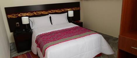 Una cama bien hecha con edredón blanco y manta estampada en rosa y rojo, cabecera de madera con diseño floral, mesita de noche con lámpara y alfombra a rayas en el suelo.