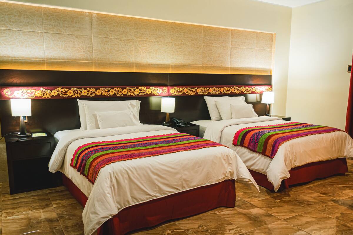A hotel room with two beds, each with a white bedspread and a colorful blanket. There are bedside tables with lamps and a decorative headboard.