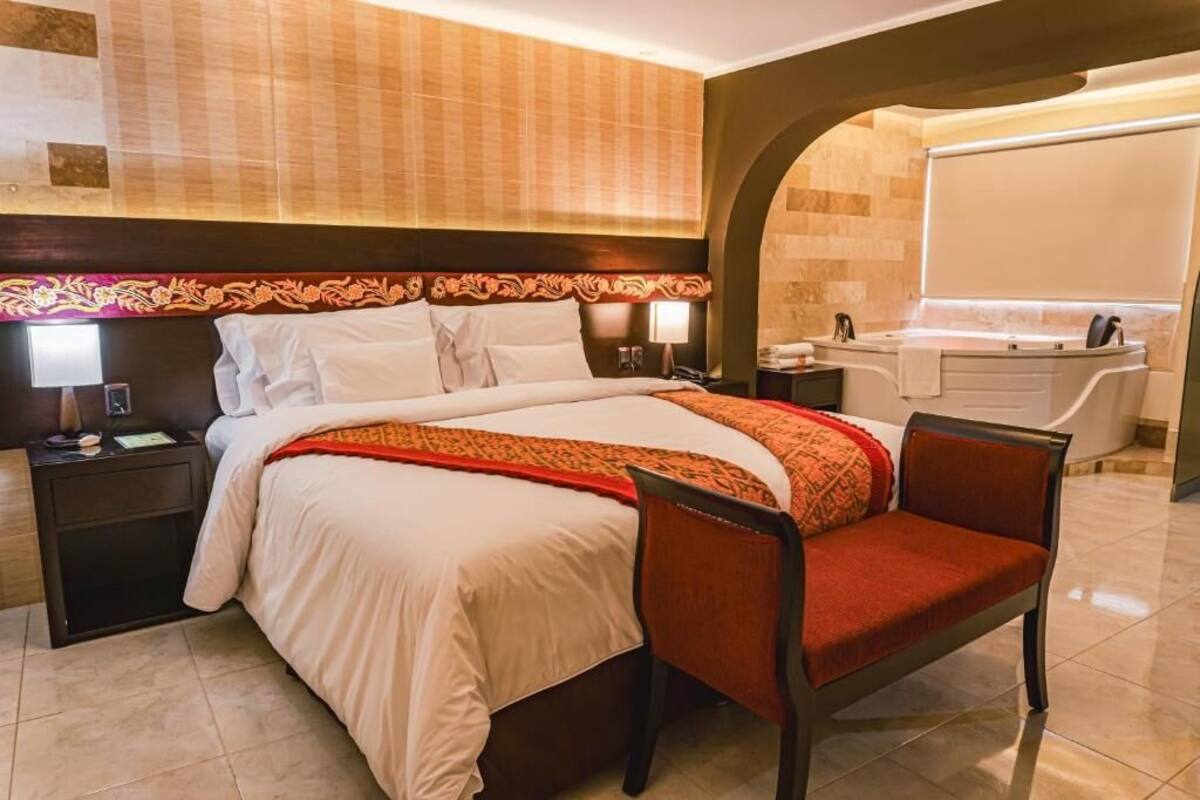 A hotel room with a bed, a chair, a nightstand, a lamp, a desk, and a bathroom with a bathtub and sink.