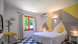 A hotel room with a checkered floor, a bed with white bedding and yellow pillows, a desk with a computer, a chair, and a window with curtains. -  Quiet family Villa in Pollensa countryside with pool,  just 3 km from town (pollensa)