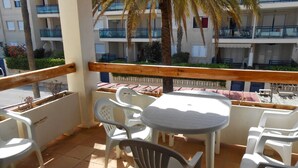 A balcony with white plastic chairs and a table, overlooking a building with balconies and palm trees.