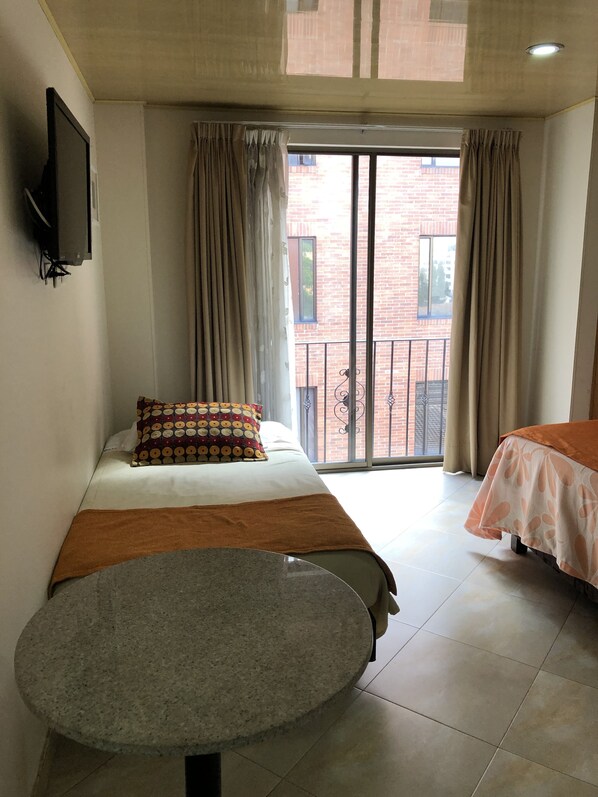 A hotel room with a bed, a small table, a TV mounted on the wall, and a view of a brick building through the window.