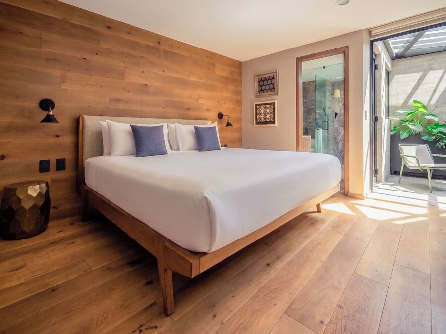 A modern bedroom with a wooden headboard, a large bed with white bedding, two wall-mounted lamps, and a glass door leading to a balcony with plants.