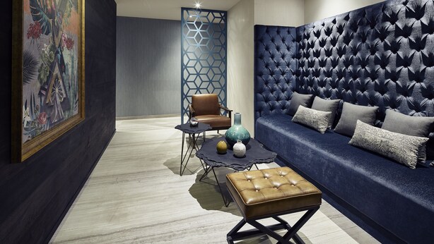 Una sala moderna con un sofá tapizado en azul oscuro, un reposapiés de cuero marrón, un jarrón decorativo y un separador geométrico.