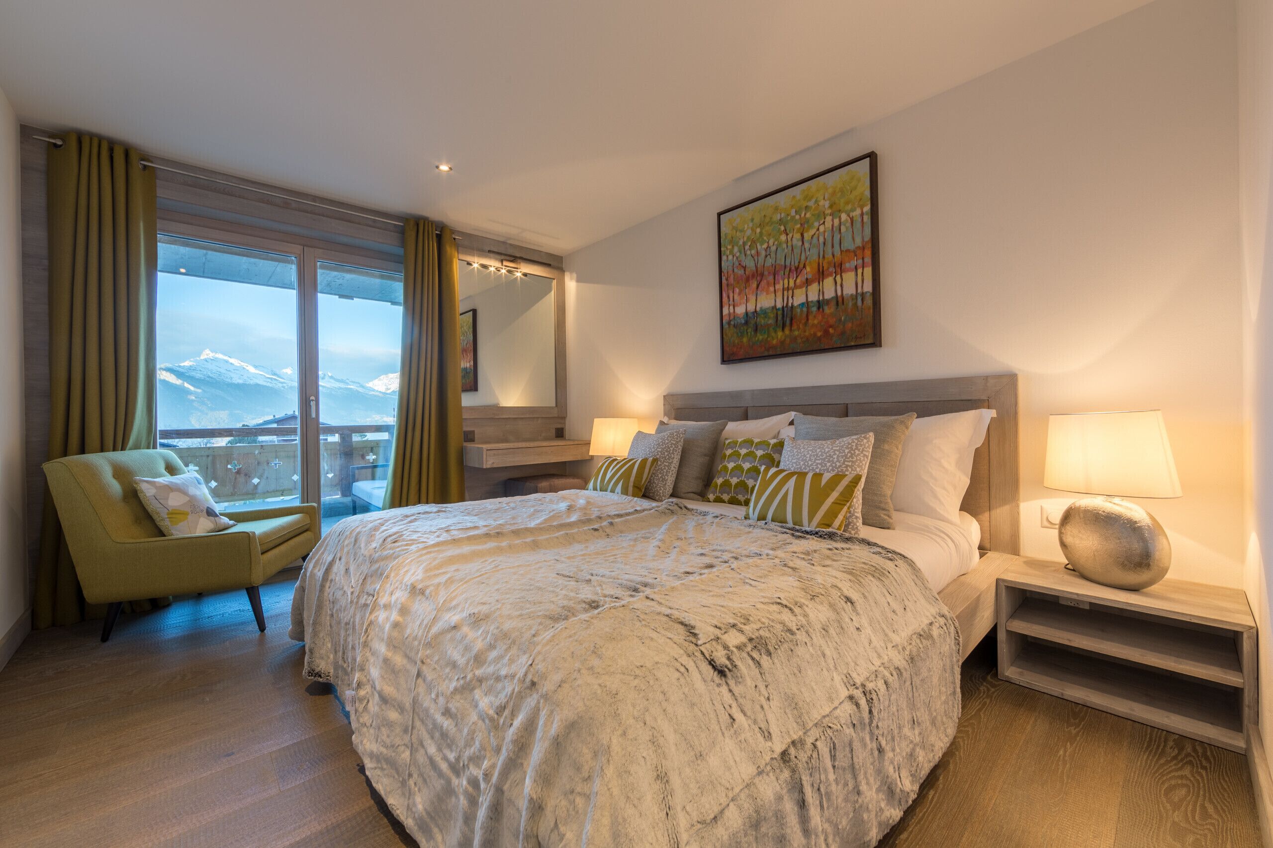 Quarto de hotel com uma cama grande, uma cadeira, uma mesinha de cabeceira, um abajur, um quadro e vista para montanhas cobertas de neve.