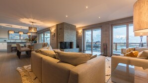 A spacious living room with a large sofa, a glass coffee table, and a fireplace. There is a dining area with a table and chairs, and large windows offering a mountain view. - La Forêt 2 - Spa access (Haute-Nendaz)