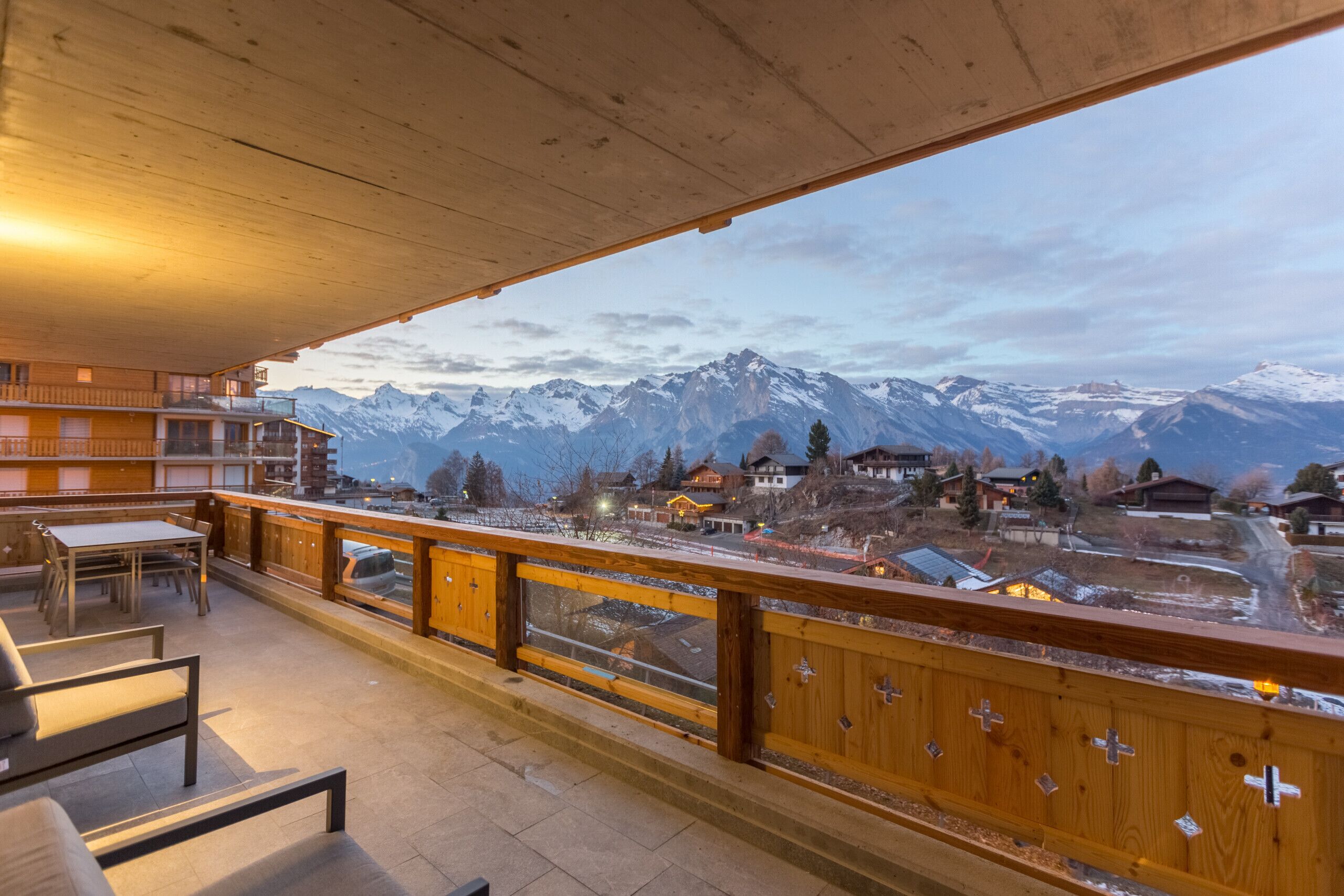 Un balcón con vistas a montañas nevadas, barandas de madera y una mesa con sillas.