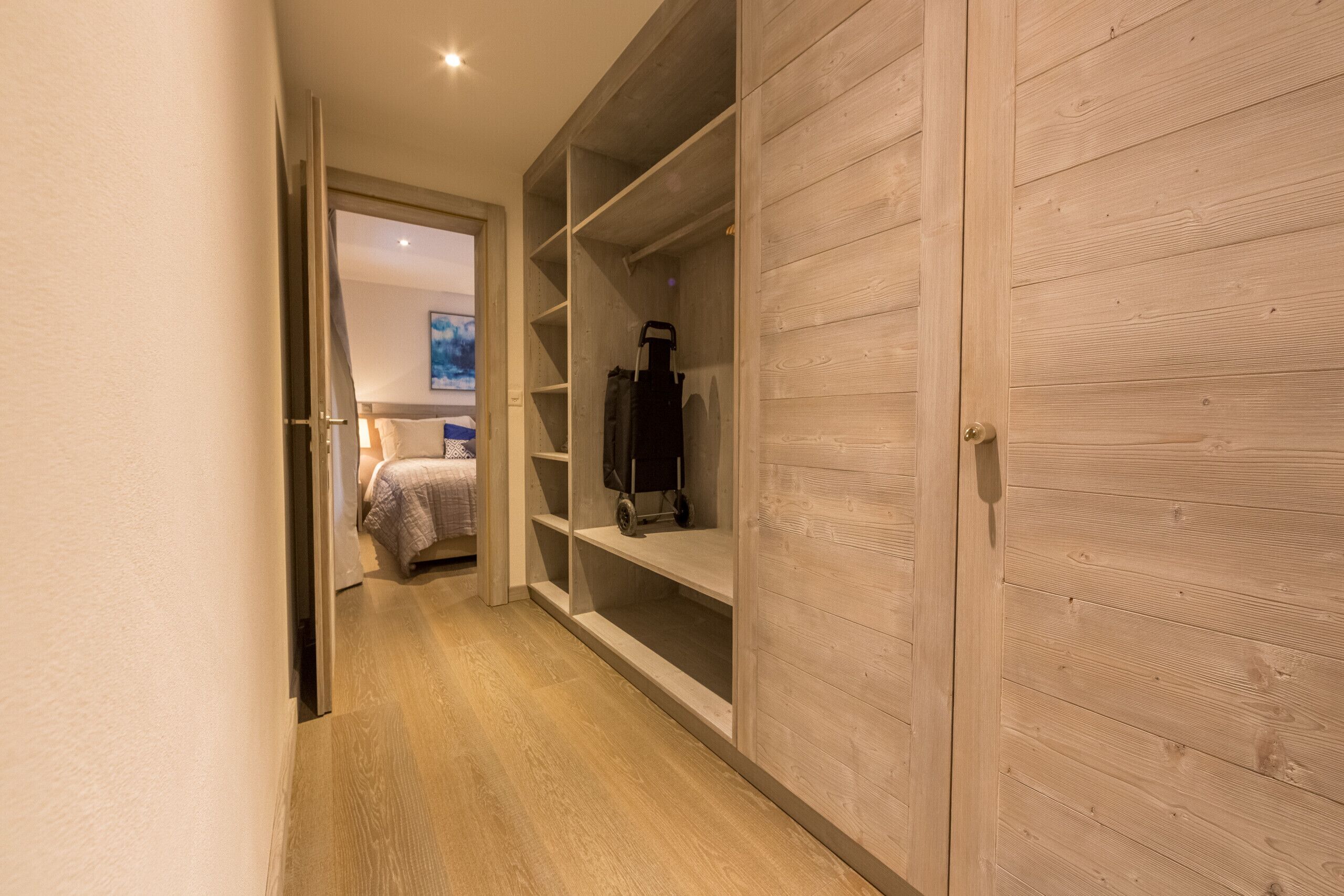 Un pasillo con un armario de madera, una cama con cabecera azul y un aspirador en un estante.