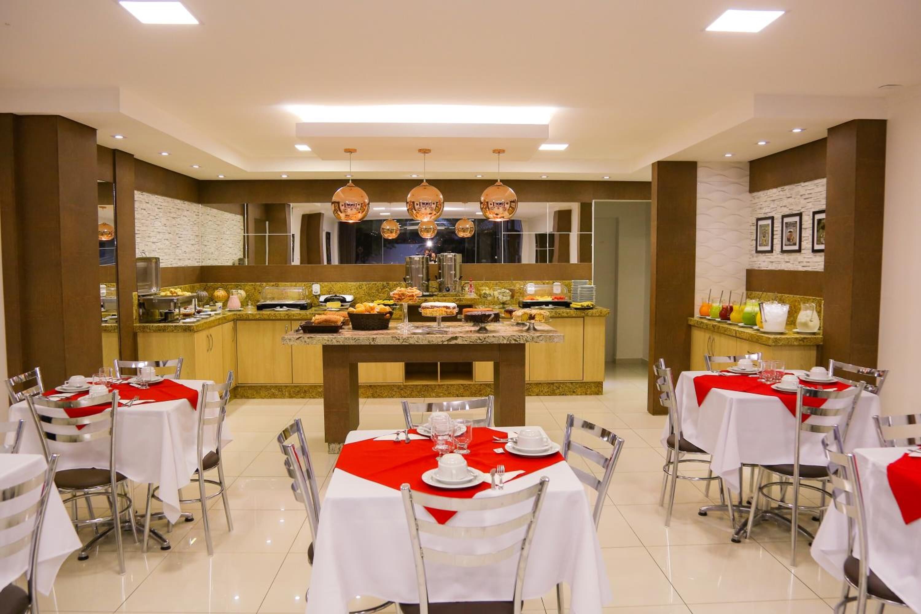 Un comedor de hotel con varias mesas preparadas para el desayuno, un bufé central con diversos alimentos, y una cocina al fondo.