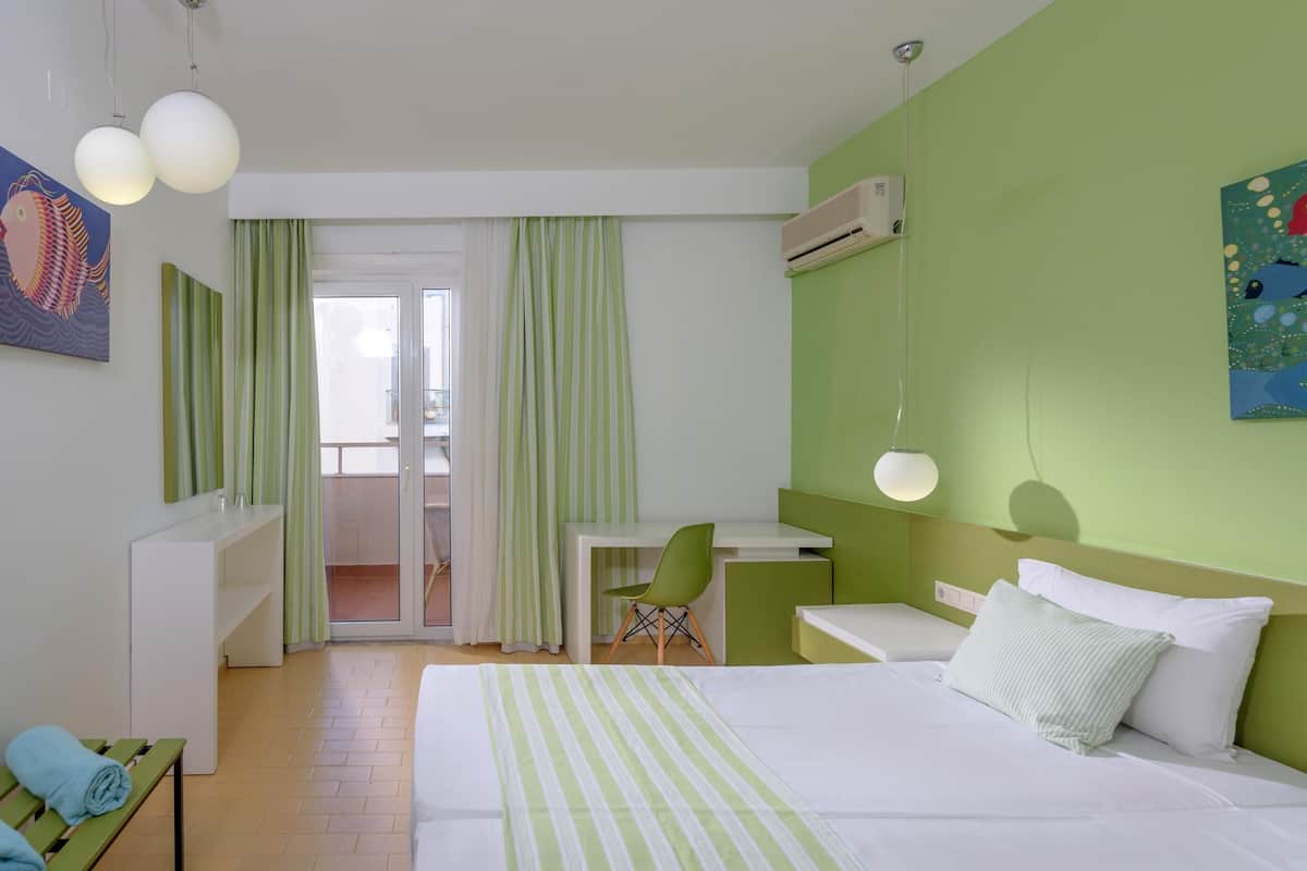 A hotel room with a bed, a desk, a chair, and a balcony with green curtains.