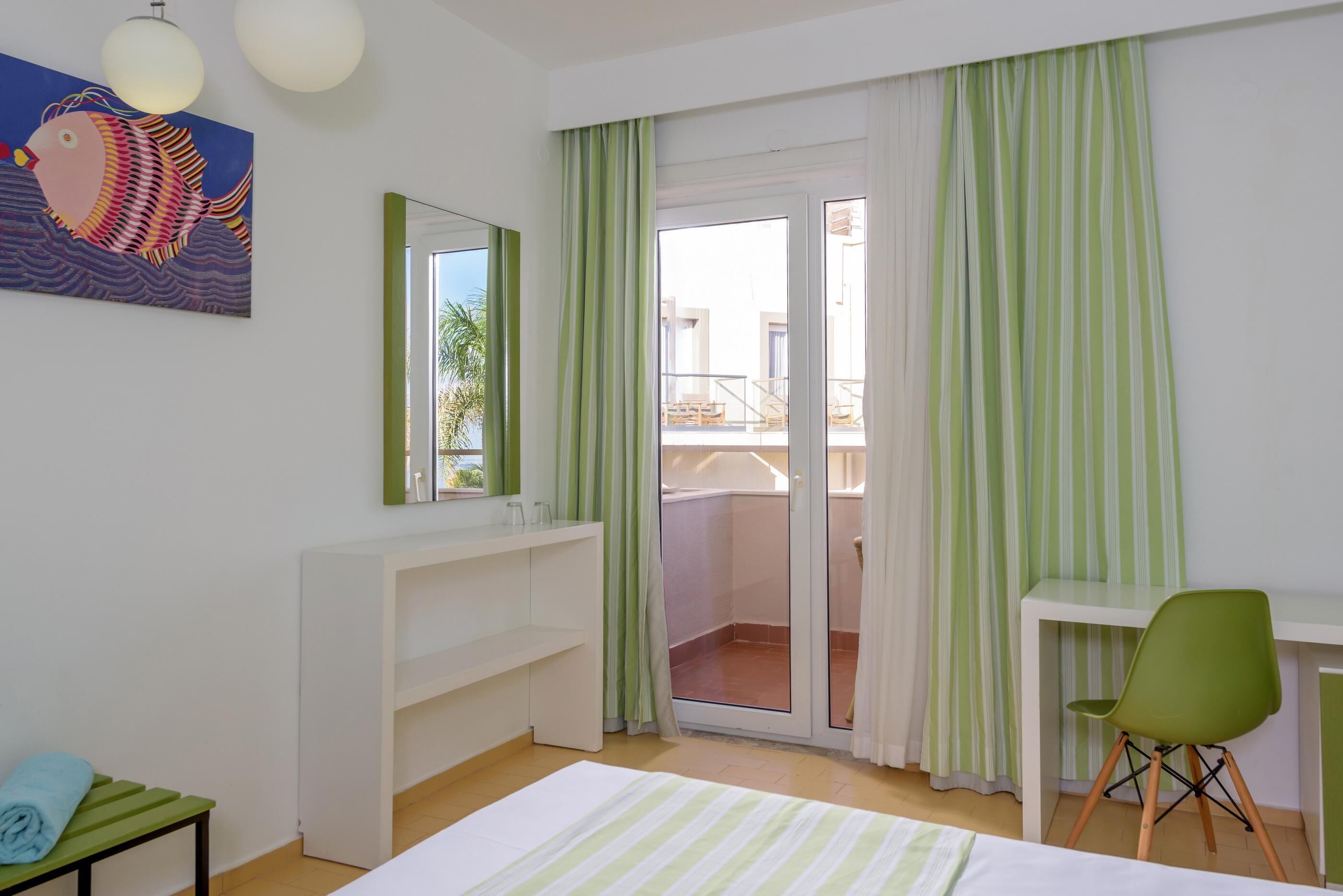 A hotel room with a bed, a desk, a chair, a bathroom with a shower, and a balcony with green curtains.