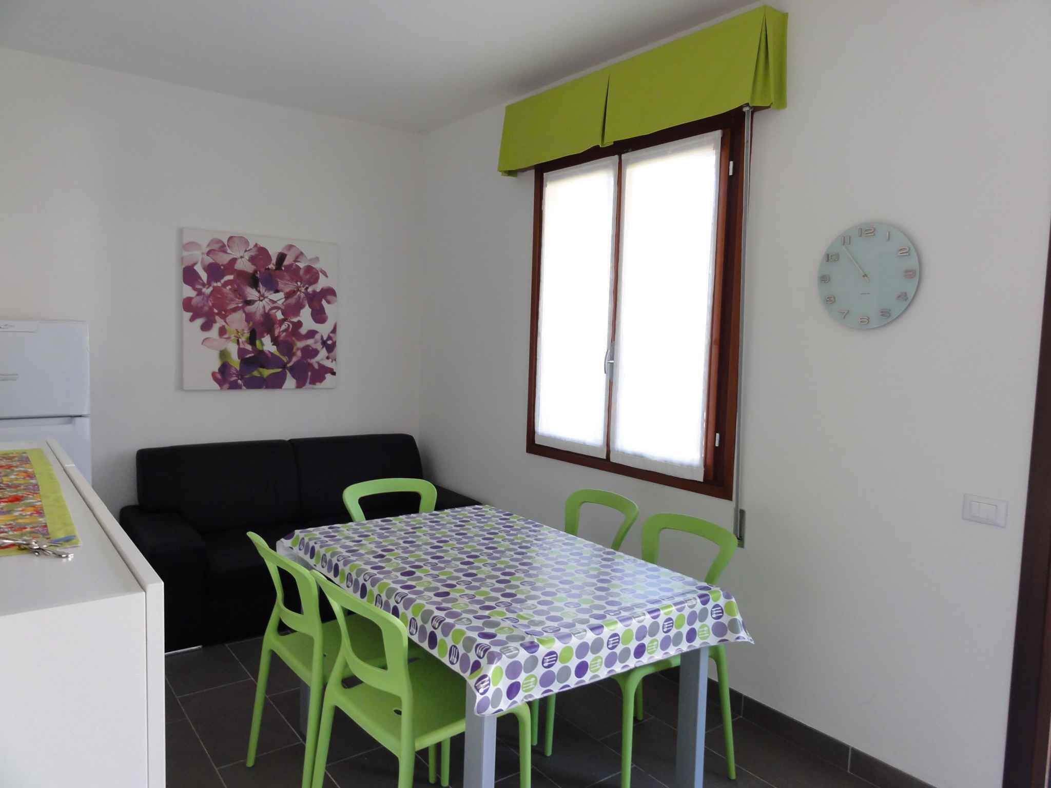 Un petit coin repas avec une table recouverte d’une nappe à pois, des chaises vertes, un canapé noir, une fenêtre agrémentée d’un lambrequin vert et une horloge au mur.