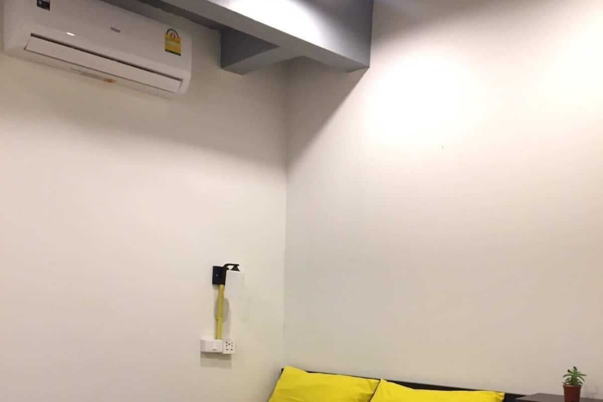 Une petite chambre comprenant un lit, un appareil de climatisation, une prise de courant et une petite plante en pot.