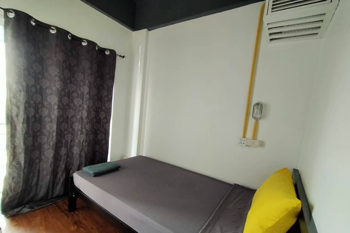 Un lit simple avec un matelas gris et un oreiller jaune, une fenêtre avec des rideaux foncés et un climatiseur fixé au mur.