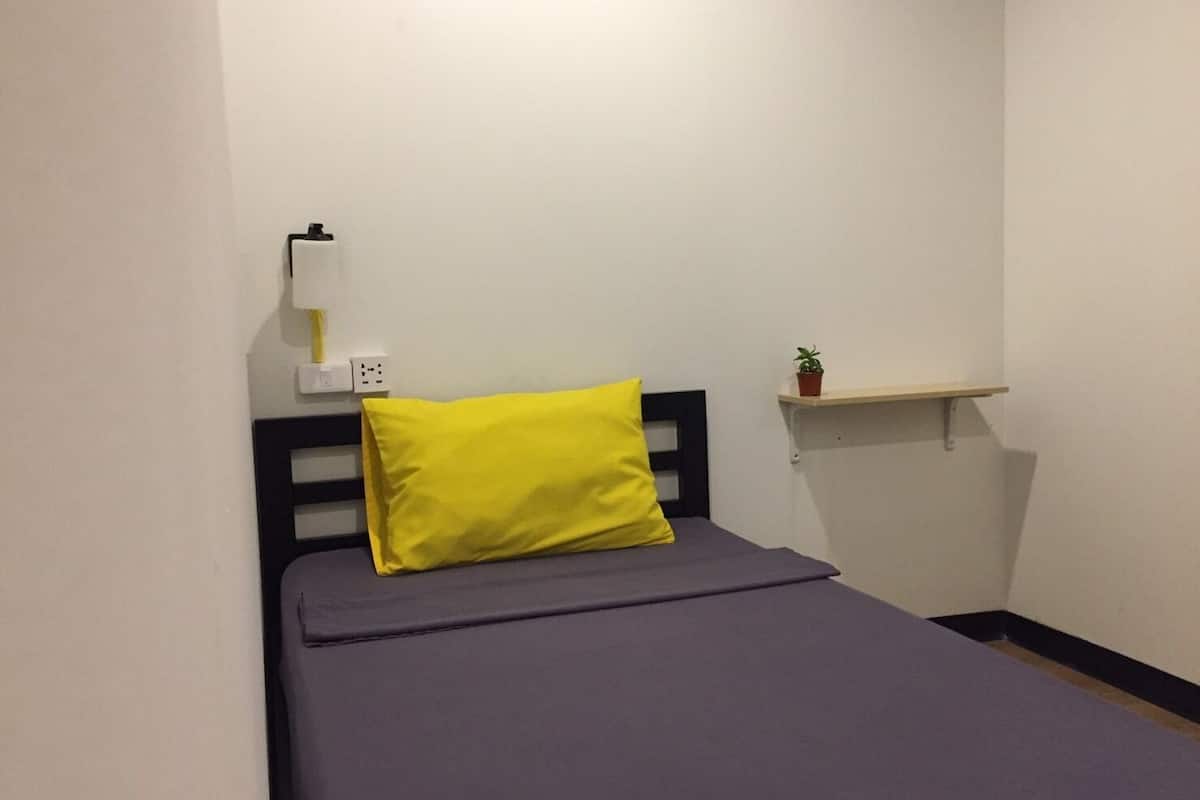 Een kleine kamer met een enkel bed, een geel kussen, een nachtkastje met een plant en een wandplank.