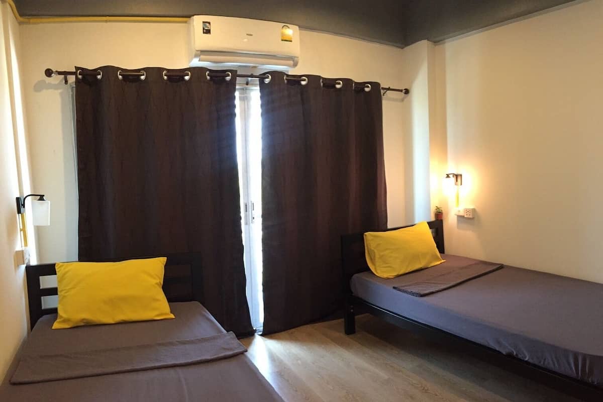 Une chambre avec deux lits, chacun étant équipé d’un oreiller et d’une couverture. Un climatiseur est installé au-dessus de la fenêtre, et le rideau est partiellement tiré.
