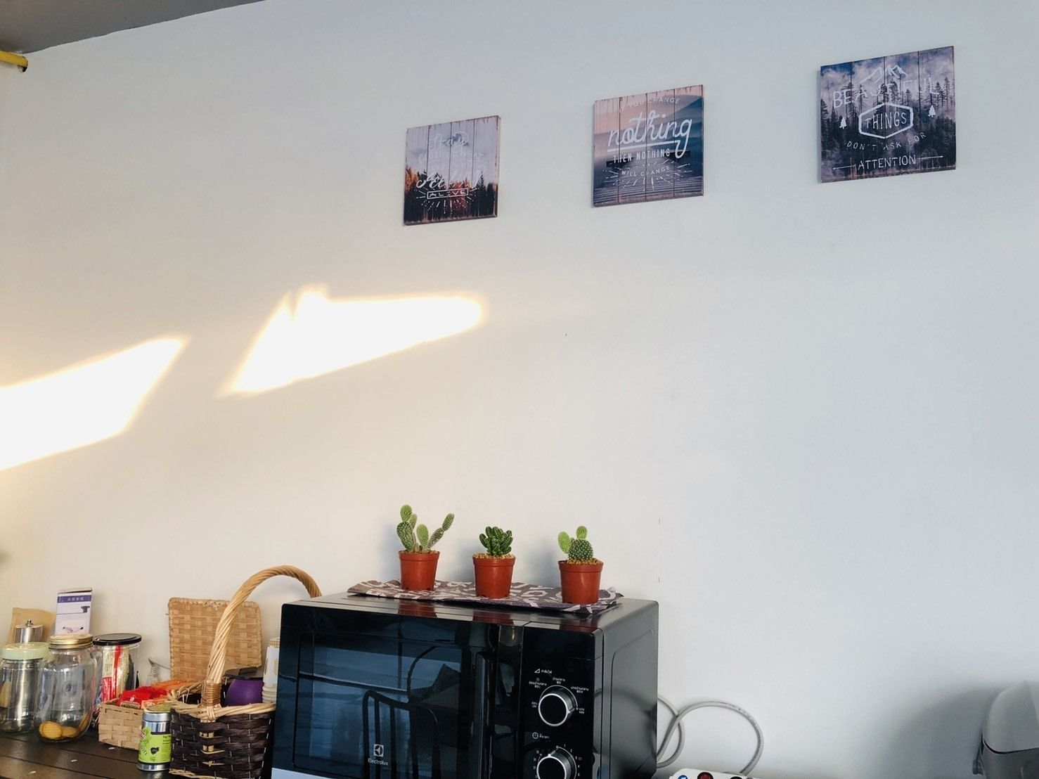 A room with a flat-screen TV on a stand, three potted cacti on top, a wicker basket, and a wall with three framed motivational posters.
