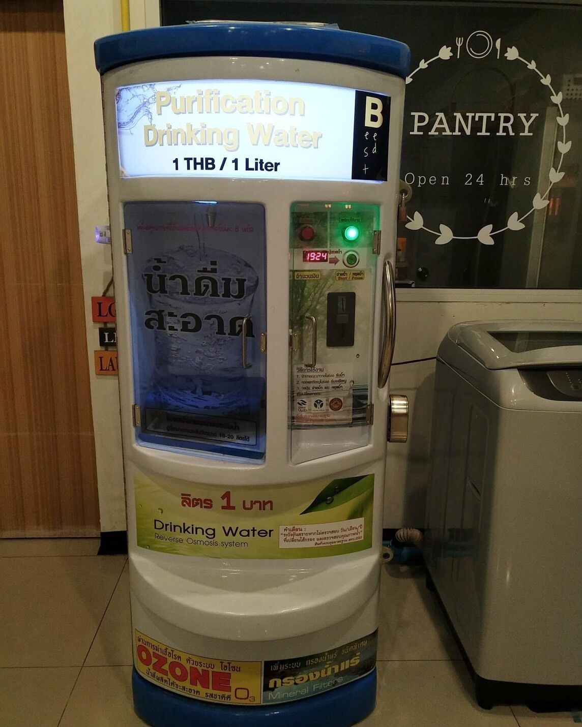 Una máquina de purificación de agua etiquetada como "1 TBH / 1 Litro" con sistema de ósmosis inversa, ubicada en una habitación con una despensa al fondo.